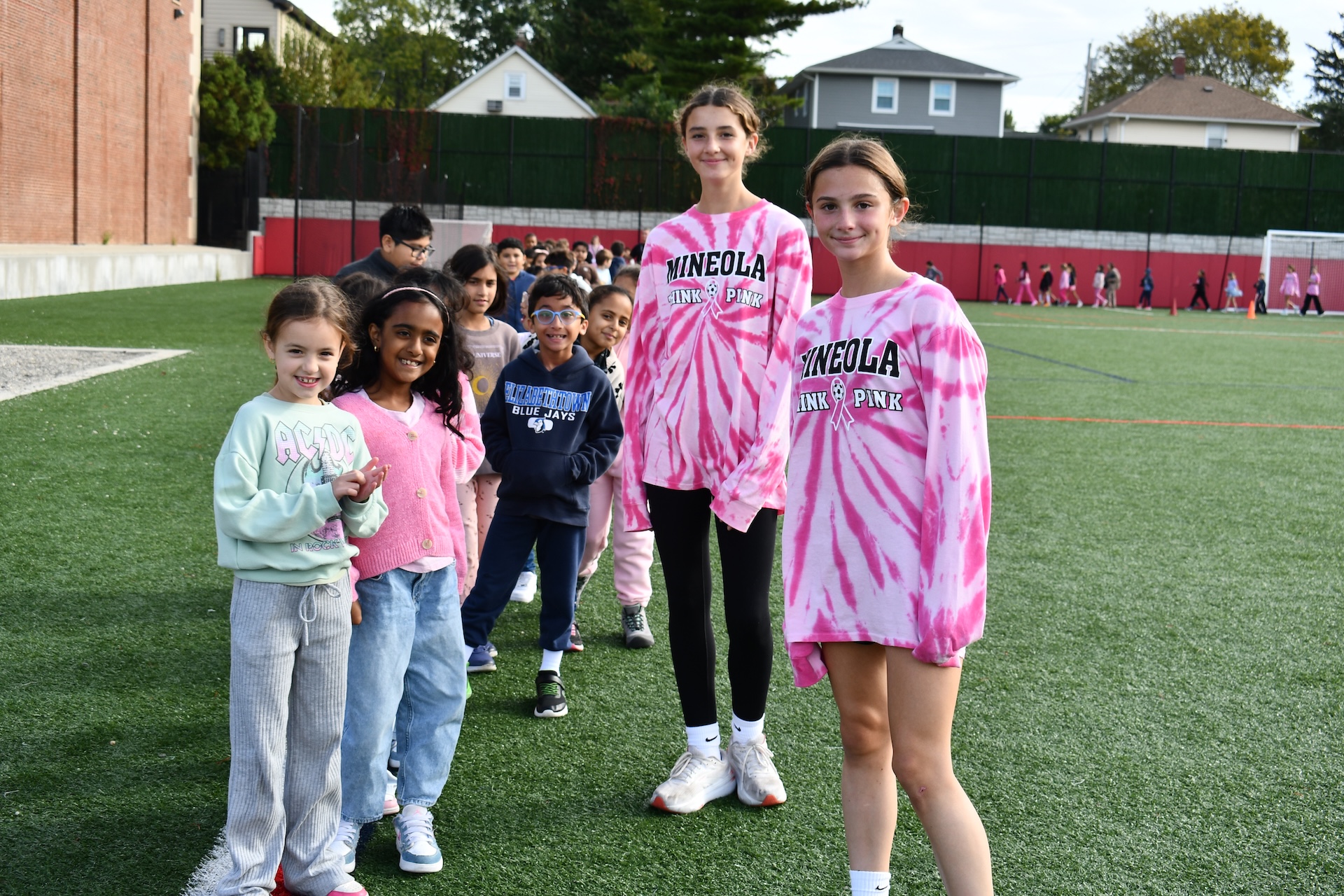 Elementary school and high school students, some wearing pink for breast cancer awareness, post on a turf field